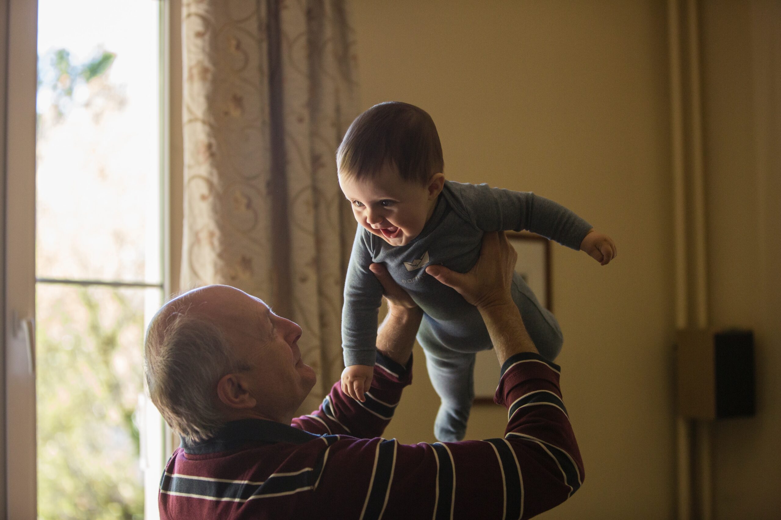 man wearing maroon, white, and blue stripe long-sleeved shirt lifting up baby wearing gray onesie