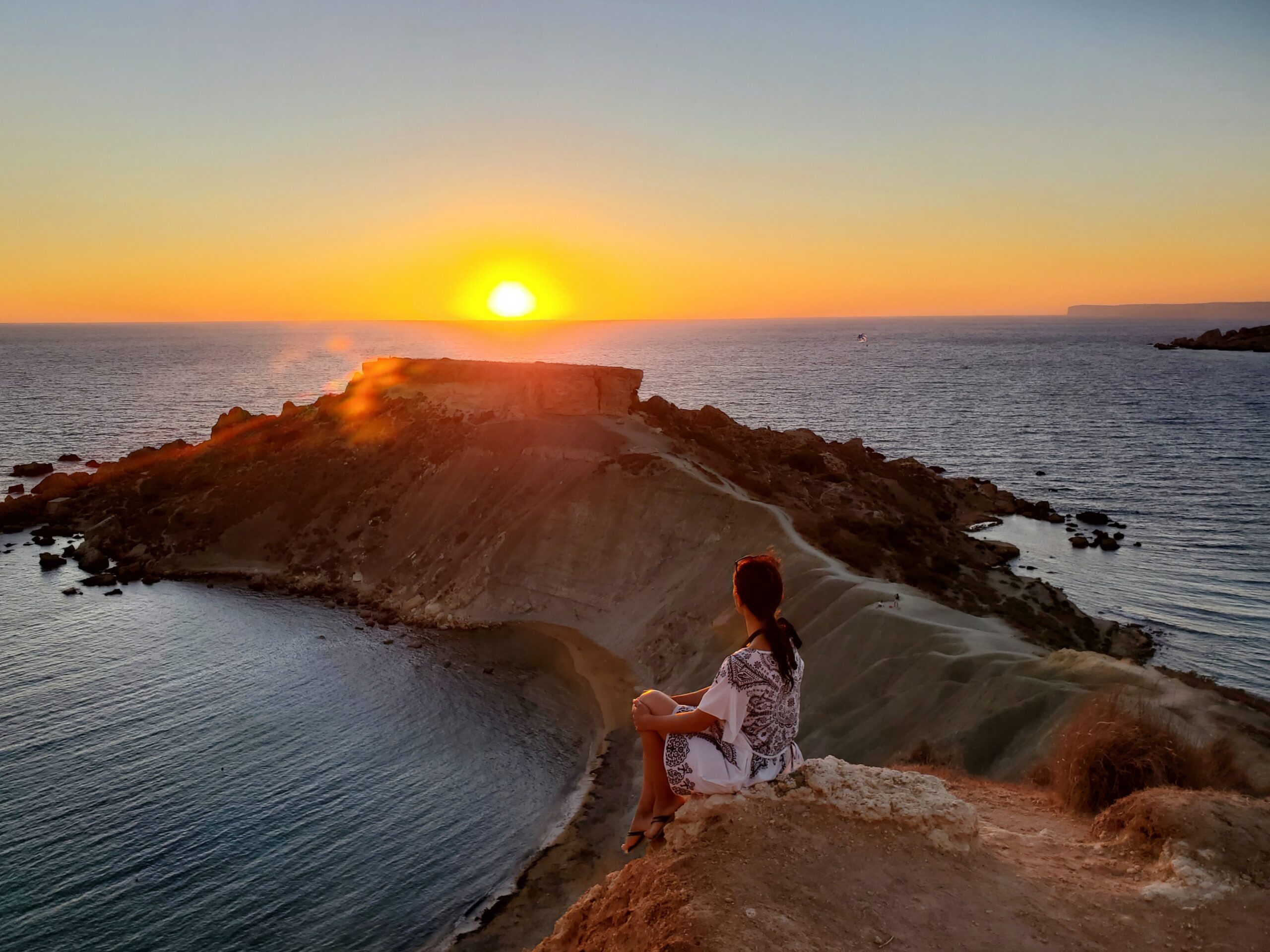 woman in white shirt standing on brown rock near body of water during sunset