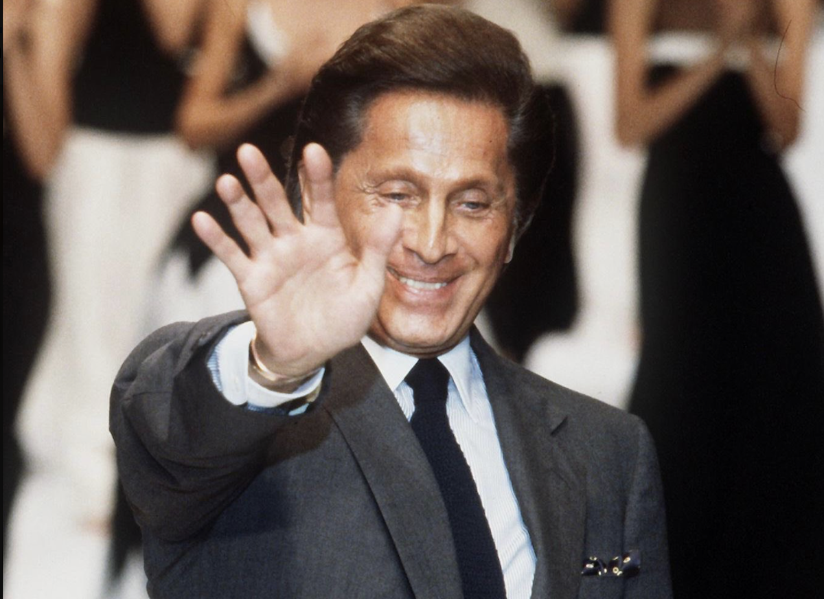 Valentino Garavani saluta il pubblico durante una sfilata, sorridente, in abito scuro