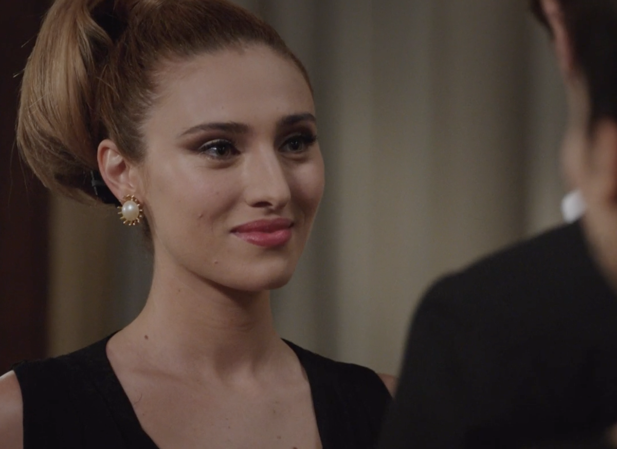 Scena elegante in interno de Il Paradiso delle Signore 10, donna con acconciatura raccolta e orecchini di perle in primo piano.